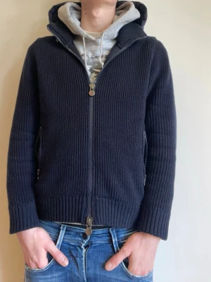 Mörkblå Cardigan från Moncler - Snygg mörkblå cardigan från Moncler med stickade ärmar och huva. Västen har dragkedja framtill, ribbade muddar och Moncler-logga på ärmen. Perfekt för lager-på-lager och ger en sportig vibe till din outfit. Kvitto finns