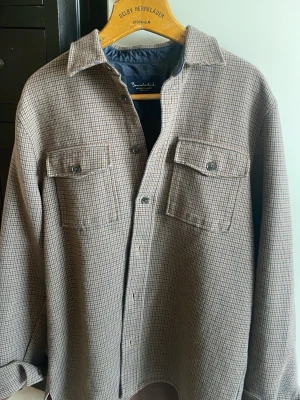 Rutig overshirt från Bondelid - Stilren overshirt från Bondelid i småmönstrat rutigt tyg i beige och brunt. Två bröstfickor med lock och knappar, klassisk krage och knäppning framtill. Perfekt lager-på-lager-plagg med mjukt innerfoder för extra komfort.