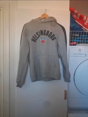 Grå Helsingborg hoodie med tryck - Snygg grå hoodie från Helsingborgs IF med stort tryck på bröstet och klubbens logga. Svarta detaljer på ärmarna och röd mudd längst ner på ärmarna. Klassisk känguruficka framtill och huva. Perfekt för dig som vill visa ditt lagstöd.