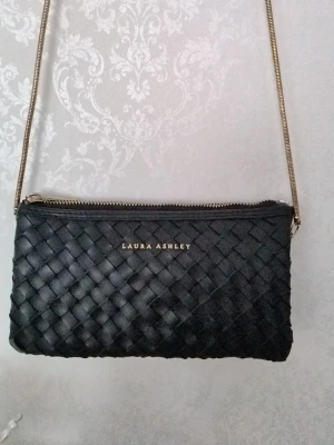 Svart axelväska Laura Ashley - Snygg svart axelväska från Laura Ashley med flätad design och guldfärgad kedjerem. Väskan har dragkedja upptill och logga i guld på framsidan. Perfekt storlek för det viktigaste och ger en stilren look till din outfit.