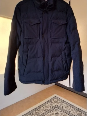 Svart  pufferjacka Tommy Hilfiger - Snygg svart  pufferjacka från Tommy Hilfiger med hög krage och fyra framfickor. Jackan har quiltad design och stängs med dragkedja och knappar. Perfekt för kalla dagar och har en klassisk streetstil.