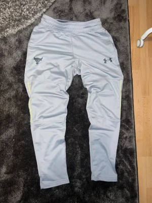 Grå mjukisbyxor från Under Armour - Säljer ett par grå mjukisbyxor från Under Armour med Project Rock-loggan och svarta detaljer. Byxorna har smal passform, resår i midjan och gula ränder längs sidorna. Perfekta för chill eller träning, riktigt sköna och stilrena.