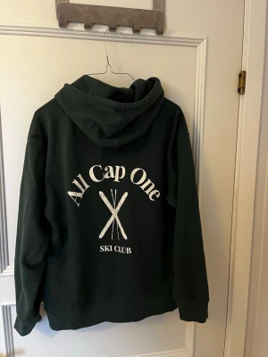 Mörkgrön hoodie All Cap One - Mörkgrön hoodie från All Cap One med stor vit text och skidmotiv på ryggen samt liten logga på bröstet. Perfekt för dig som gillar clean och trendig stil.