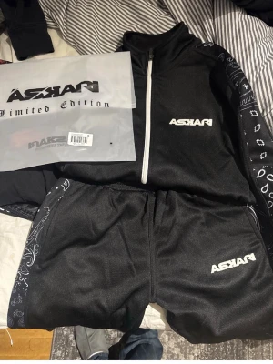 Svart tracksuit från Askari - Säljer en svart tracksuit från Askari Limited Edition med vita detaljer och dragkedja framtill. Setet har vita logotyper på både jacka och byxor samt mönstrade bandana-detaljer längs ärmar och ben. Perfekt för en sportig och avslappnad stil. Mycket bra skick som nytt. LIMITED EDITION 