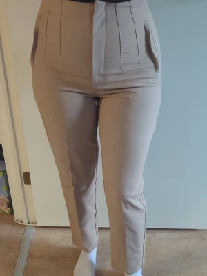 Kostymbyxor med hög midja - Snygga beige kostymbyxor med hög midja och pressveck framtill. Byxorna har raka ben, diskreta fickor och stängs med dragkedja och hake. Materialet är slätt och ger en stilren look, perfekt till en dressad outfit.