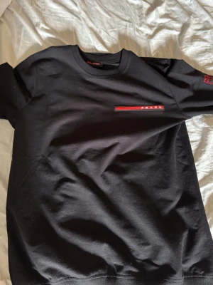 Svart sweatshirt från Prada - Snygg svart sweatshirt från Prada med klassisk röd logga på bröstet och ribbade muddar vid hals, ärmslut och nederkant. Perfekt för dig som gillar stilrena designer och exklusiva märken. Materialet känns mjukt och bekvämt mot huden.