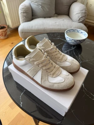 Beige och vita sneakers med mockadetaljer - Snygga sneakers i beige mocka och vitt skinn med klassisk gummisula. Lågt skaft och stilren design som passar till det mesta. Perfekta för dig som gillar minimalistisk och tidlös stil.