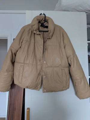Beige pufferjacka  - Säljer en beige pufferjacka med hög krage och tryckknappar framtill. Jackan har två stora fickor på sidorna och är tillverkad i ett glansigt material som liknar konstläder. Har lite slitage på kragen men syns inte när man har den på sig.