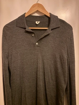 Mörkgrå Arket långärmad tröja med krage - Snygg mörkgrå långärmad tröja från Arket med klassisk krage och tre knappar vid halsen. Tillverkad i mjukt och tunt material som känns skönt mot huden. Perfekt för en stilren och avslappnad look.