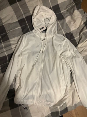 Vit vindjacka från Risky - Snygg vit vindjacka från Risky med dragkedja framtill, huva med dragsko och två sidofickor. Jackan har en minimalistisk design och är tillverkad i ett lätt, vindtätt material. Perfekt för blåsiga dagar och enkel att matcha med olika outfits. 