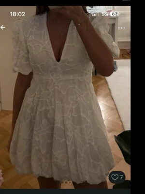 Vit kort klänning med puffärm - Supersöt vit kortklänning med puffärmar och djup v-ringning. Klänningen har broderade blommor över hela tyget och en volangig kjol med ballongformad nederkant. Perfekt för sommarens alla tillfällen!