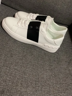 Valentino vita sneakers med svart band 43 - Snygga vita sneakers från Valentino med ett brett svart band över vristen och coola nitar på hälen. Skorna är i slätt läder och har vita skosnören. Perfekta för dig som gillar stilrena men ändå unika detaljer.