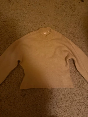 Beige stickad tröja med hög krage - En enkel och stilren beige stickad tröja med hög krage och långa ärmar. Tröjan har en rak passform och ribbade muddar vid ärmslut och nederkant. Perfekt att matcha med jeans eller kjol för en avslappnad look.