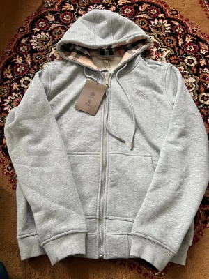 Grå zip hoodie från Burberry - Snygg grå hoodie från Burberry med dragkedja framtill och klassiskt rutigt foder i huvan. Tröjan har ribbade muddar, två fickor och justerbar dragsko i huvan. Perfekt för en chill och stilren look. Materialet är mjukt och skönt, troligen bomullsblandning.