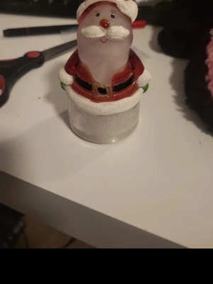 Dekorativ tomtefigur i glas - Säljer en söt tomtefigur i glas med röd jacka, svart bälte och vita detaljer. Figurer har ett glatt ansikte, vit luva och skägg samt gröna vantar. Perfekt som juldekoration på hyllan eller bordet.