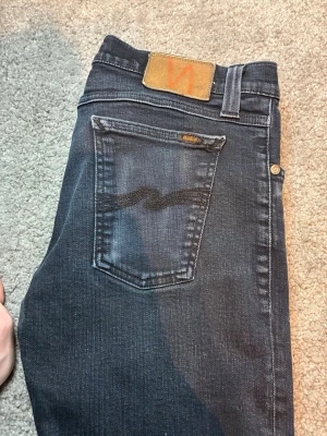 Mörkblå jeans från Nudie Jeans - Säljer ett par mörkblå jeans från Nudie Jeans med klassisk femficksmodell och snygga kontrastsömmar. Bakfickan har Nudie Jeans karakteristiska vågmönster och läderpatch i midjan. Perfekta för dig som gillar stilrena och tidlösa jeans.