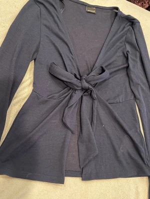Mörkblå knytblus med långa ärmar - Trendig mörkblå topp från Gina tricot med långa ärmar och V-ringning. Toppen har en snygg knytning framtill som ger en cool och avslappnad look. Tillverkad i ett mjukt, stretchigt material som sitter skönt på kroppen. Perfekt till jeans eller kjol.