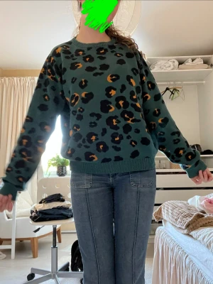 Grön leopardmönstrad stickad tröja - Säljer en grön stickad tröja med leopardmönster i svart och orange. Tröjan har rund halsringning, ribbade muddar vid ärmslut och nederkant samt en avslappnad passform. Perfekt för dig som vill sticka ut lite extra med ett djurmönster. Ifrån dedicated vilket gör den mycket miljövänlig och bra kvalitet