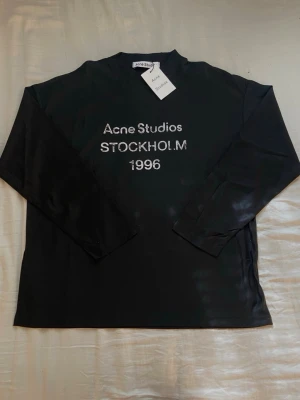 Svart långärmad topp Acne Studios - Svart långärmad topp från Acne Studios med tryckt text 'Acne Studios STOCKHOLM 1996' framtill. Enkel och stilren design med rund hals. Perfekt för dig som gillar minimalistisk och trendig stil.