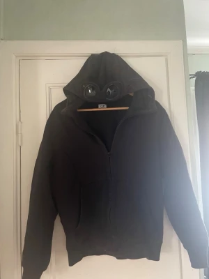 Svart hoodie med glasögon i huvan - Säljer en svart hoodie från C.P. Company med dragkedja framtill och deras ikoniska glasögon i huvan. Hoodien har känguruficka och ribbade muddar vid ärmar och nederkant. Perfekt för dig som vill sticka ut med en unik detalj. Är i använt skick!!!!!!!!!!