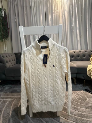 Vit tröja Polo Ralph Lauren - Säljer en snygg vit kabelstickad tröja från Polo Ralph Lauren med hög krage och broderad logotyp på bröstet. Tröjan har långa ärmar och ribbade muddar vid ärmslut och nederkant. Perfekt för dig som gillar klassisk och stilren design.                                         PASSAR  bra till folk med storlek S-M 