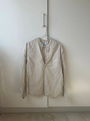 Ljusbeige henley-skjorta i bomull - Ljusbeige henley-skjorta med lång ärm och klassisk knäppning vid halsen. Skjortan är tillverkad i mjuk bomull och har en enkel, stilren design utan mönster. Perfekt för dig som gillar en avslappnad och modern look.