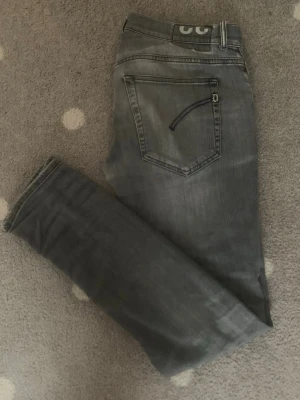 Dondup jeans grå - Snygga grå slim fit jeans från Dondup med slitningar och tvättade detaljer. Klassisk femficksmodell med dragkedja och knapp. Bakfickan har en unik detalj med broderad båge. Perfekta för en avslappnad och trendig look. Har 2 små hål i varje knä del av jeansen, men det har sytts ihop. 
