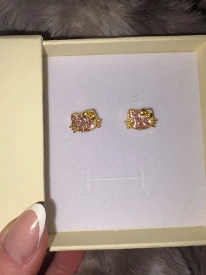 Hello Kitty örhängen - Söta örhängen i form av Hello Kitty-huvuden med rosa glitter och guldfärgade detaljer. Perfekta för dig som gillar lekfulla accessoarer med en touch av nostalgi. Kommer i en fin ask.