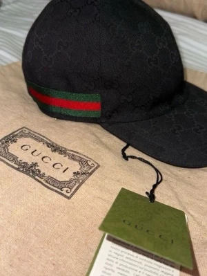 Svart GG-keps  - Svart keps med diskret GG-logomönster över hela kepsen. Klassisk böjd skärm och justerbar passform. Sidorna har gröna och röda ränder. Tillverkad i canvasmaterial.