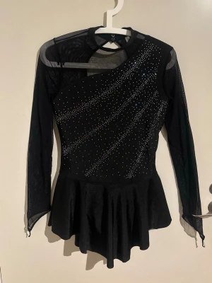 Konståkningsklänning  - Klänningen har genomskinliga långa ärmar i mesh och en stilren design. Den har även små gummiband vid ärmsluten som man sätter runt fingret, så att ärmarna håller sig på plats under åkning – perfekt för träning och tävling.  Den är aldrig använd men utan prislapp