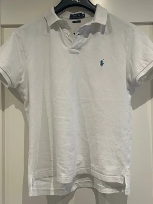 Vit pikétröja från Polo Ralph Lauren - Klassisk vit pikétröja från Polo Ralph Lauren med blå broderad logga på bröstet. Tröjan har korta ärmar, krage och knappar vid halsen. Slim fit-modell i mjuk bomull som passar perfekt till jeans eller shorts.