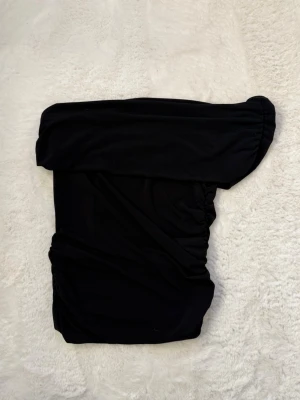 Svart offshoulder topp - Trendig svart offshoulder topp med en axel och rynkade detaljer. Perfekt för dig som vill ha en stilren och modern look. Toppens material känns mjukt och följsamt mot huden. .