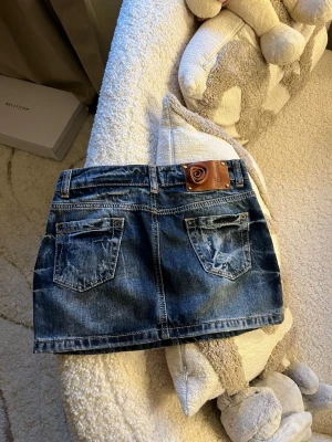 Jeanskjol från Red Valentino - Snygg kort jeanskjol från Red Valentino med två bakfickor och ett brunt lädermärke med rosdetalj bak i midjan. Klassisk blå denim med slitningar och kontrastsömmar. Perfekt för en trendig och avslappnad look.