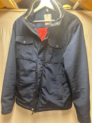 Marinblå jacka från J.Lindeberg - Stilren marinblå jacka från J.Lindeberg med quiltat rött innerfoder. Jackan har flera fickor framtill, dragkedja och knappstängning samt en hög krage med randig detalj. Materialet är slätt och vattenavvisande, perfekt för kyliga dagar.