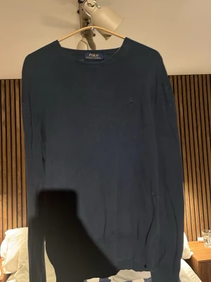 Mörkblå tröja från Polo Ralph Lauren - Klassisk mörkblå tröja från Polo Ralph Lauren med rund halsringning och diskret logga på bröstet. Tröjan har lång ärm och är tillverkad i ett mjukt material som passar perfekt till många olika stilar.