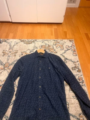 Mörkblå mönstrad skjorta från Morris  - Snygg mörkblå skjorta med diskret blommigt mönster och klassisk krage. Skjortan har långa ärmar och knäppning framtill. Perfekt för dig som vill ha en stilren men ändå unik look. Storleken är i S men enligt mig själv ligger den mellan S och M. Priset kan pratas om och jag kan även skicka lite fler bilder.