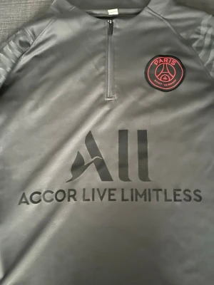 Grå PSG träningströja från Nike - Snygg grå träningströja från Nike med Paris Saint-Germain-logga på bröstet och röd Nike Swoosh på ärmen. Tröjan har halv dragkedja framtill och svart tryck med 'Accor Live Limitless' på magen. Tillverkad i ett sportigt, lätt material.