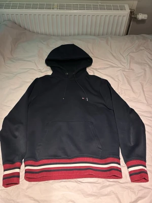 Tommy Hilfiger Hoddie  - Snygg marinblå hoodie från Tommy Hilfiger med huva och dragsko. Har en klassisk känguruficka framtill och ribbade muddar i rött, vitt och marinblått vid ärmslut och nederkant. Liten logga på bröstet. Perfekt för en avslappnad och stilren look.