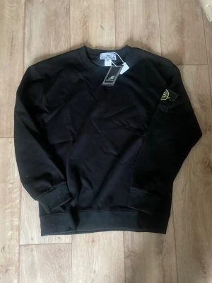 Svart sweatshirt från Stone Island - Säljer en svart sweatshirt från Stone Island med klassisk rund halsringning och ribbade muddar. Tröjan har den ikoniska Stone Island-loggan på ärmen och är tillverkad i mjuk bomull. Perfekt för en clean och stilren look.