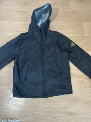 Svart vindjacka från Stone Island - 1,1 Snygg svart vindjacka från Stone Island med huva och dragkedja framtill. Jackan har två snedställda fickor med dragkedja och den klassiska Stone Island-loggan på vänster ärm. Tillverkad i lätt, vattenavvisande material som passar perfekt för blåsiga dagar.