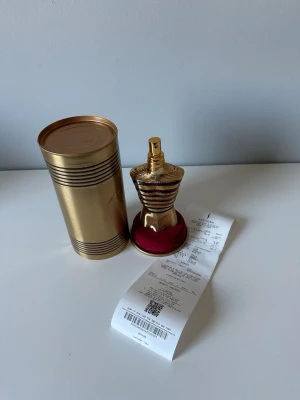 Jean Paul Gaultier Le Male Parfym - Säljer en Jean Paul Gaultier Le Male parfym med en unik flaska formad som en torso i guld och röd färg. Flaskan är tillverkad i metall och kommer med en matchande guldfärgad cylinderformad förpackning. Perfekt för dig som vill ha något exklusivt på hyllan.