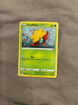 Pokémonkort Gossifleur - Säljer ett Pokémonkort med Gossifleur-motivet. Kortet har en grön bakgrund med blommor och Gossifleur i gul och röd färg. Kortet är tillverkat i papper och har en klassisk rektangulär form. Perfekt för samlare eller Pokémon-fans.
