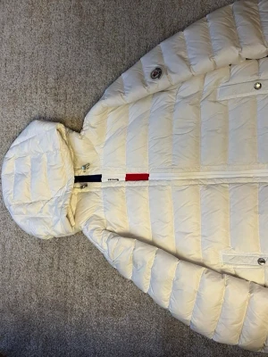 Moncler Puffer Jacket - Säljer en snygg vit dunjacka från Moncler med huva och dragkedja. Jackan har Moncler-logga på ärmen, två fickor med knapp framtill och coola detaljer i rött och blått vid dragkedjan. Perfekt för alla väder och riktigt stilren look.