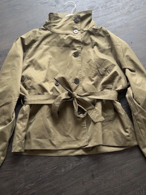 Kort grön trenchcoat med knytskärp - Grön trenchcoat med hög krage, dubbelknäppning och knytskärp i midjan. 