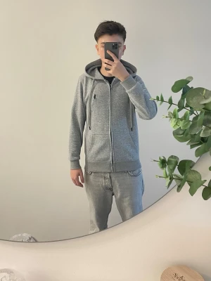 Polo Ralph Lauren Zip-Up Hoodie – Grå Heather - Säljer nu en klassisk grå zip-hoodie från Polo Ralph Lauren. Tröjan har den ikoniska svarta hästen broderad på bröstet och en mjuk, skön insida. Perfekt passform som funkar till både jeans och mjukisbyxor. 