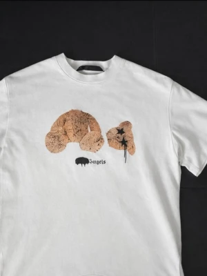 Vit t-shirt Palm Angels björntryck - Vit t-shirt från Palm Angels med ett stort tryck av en trasig nallebjörn på bröstet och svart text under. Klassisk rund halsringning och korta ärmar. Perfekt för dig som gillar streetwear och unika prints.