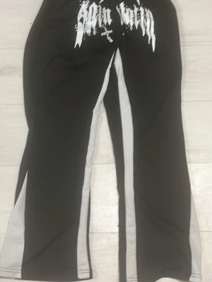 Svarta leggings med vit text - Säljer ett par svarta leggings med vita paneler längs benen och vit tryckt text framtill i en edgy stil. De har utsvängda ben och är tillverkade i ett stretchigt material som sitter skönt. Perfekta för dig som gillar streetwear och vill sticka ut.