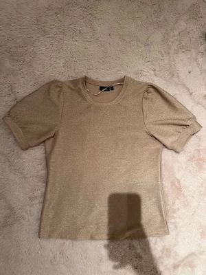 Glittrig guld topp med puffärm - Säljer en glittrig guld topp från H&M med puffärmar och rund halsringning. Toppen har en figurnära passform och korta ärmar. Perfekt för dig som vill sticka ut lite extra med en snygg metallic-look.