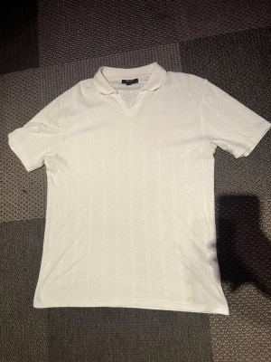 Vit kortärmad t-shirt från CAV - Säljer en stilren vit t-shirt från CAV med krage och v-ringning. T-shirten har ett diskret mönster i tyget och är kortärmad, perfekt för en clean och enkel look. Materialet känns mjukt och bekvämt, troligtvis bomull eller polyester.