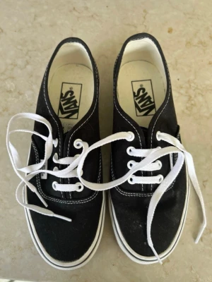Svarta Vans sneakers med vita detaljer - Klassiska svarta Vans sneakers med vit sula och vita skosnören. Skorna har låg profil, canvasmaterial och tydliga vita sömmar. Vans-logga på sidan och innersulan. Perfekta för en avslappnad och stilren look.